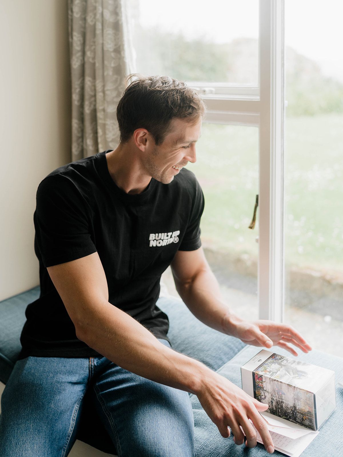 Person in black t-shirt sits by window examining a box with printed imagery.