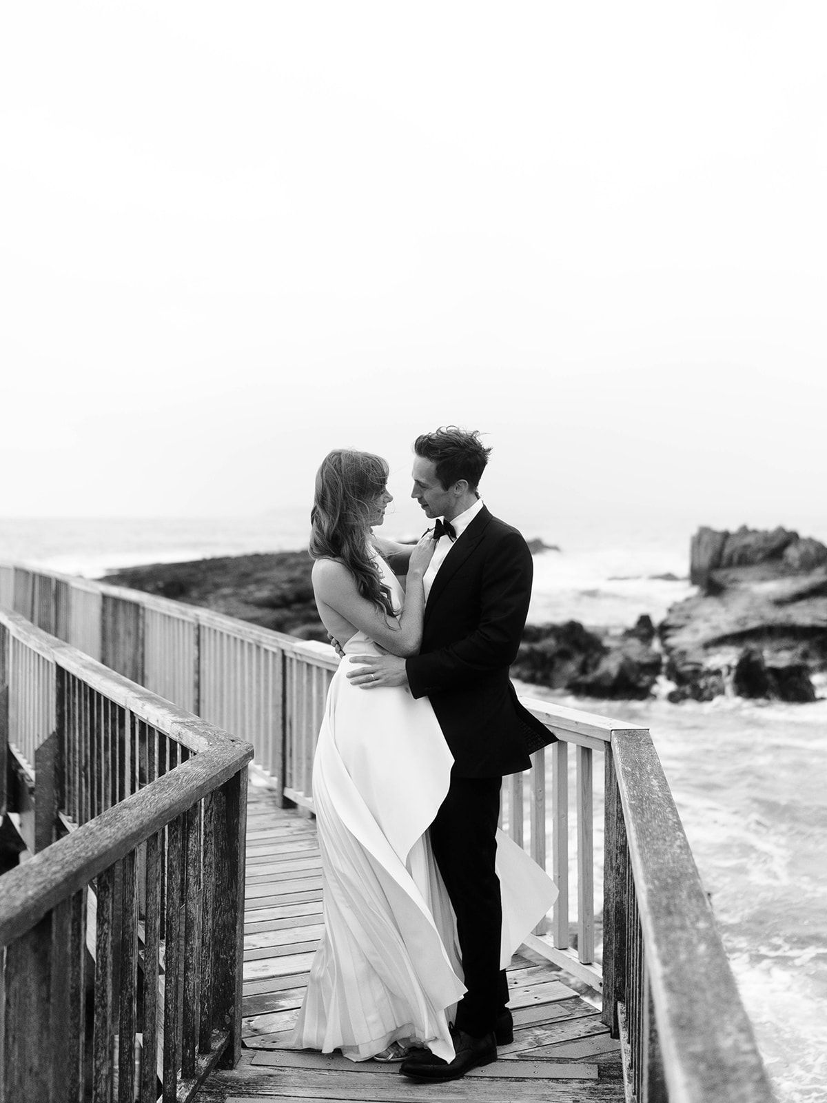 A couple in formal attire shares a tender moment on a seaside boardwalk.