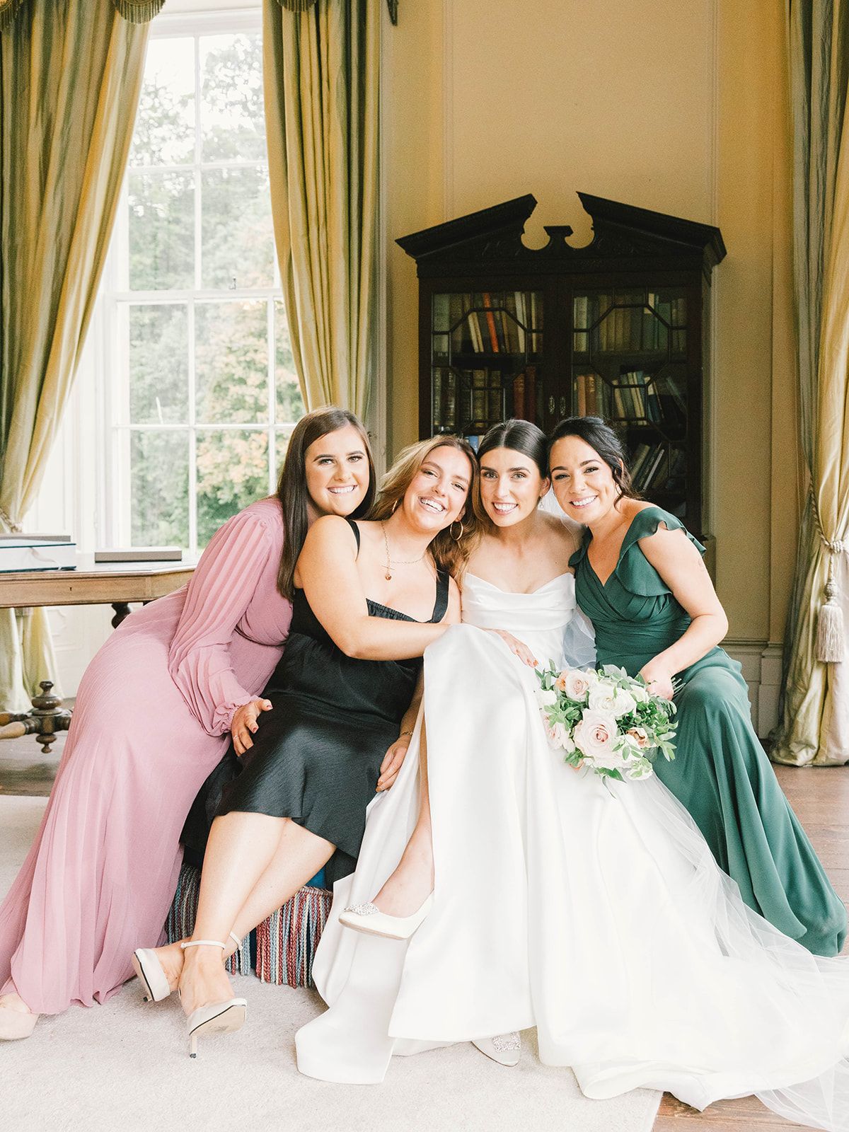 A bridal party in colorful dresses poses together in an elegant room with yellow curtains and a vintage bookshelf.