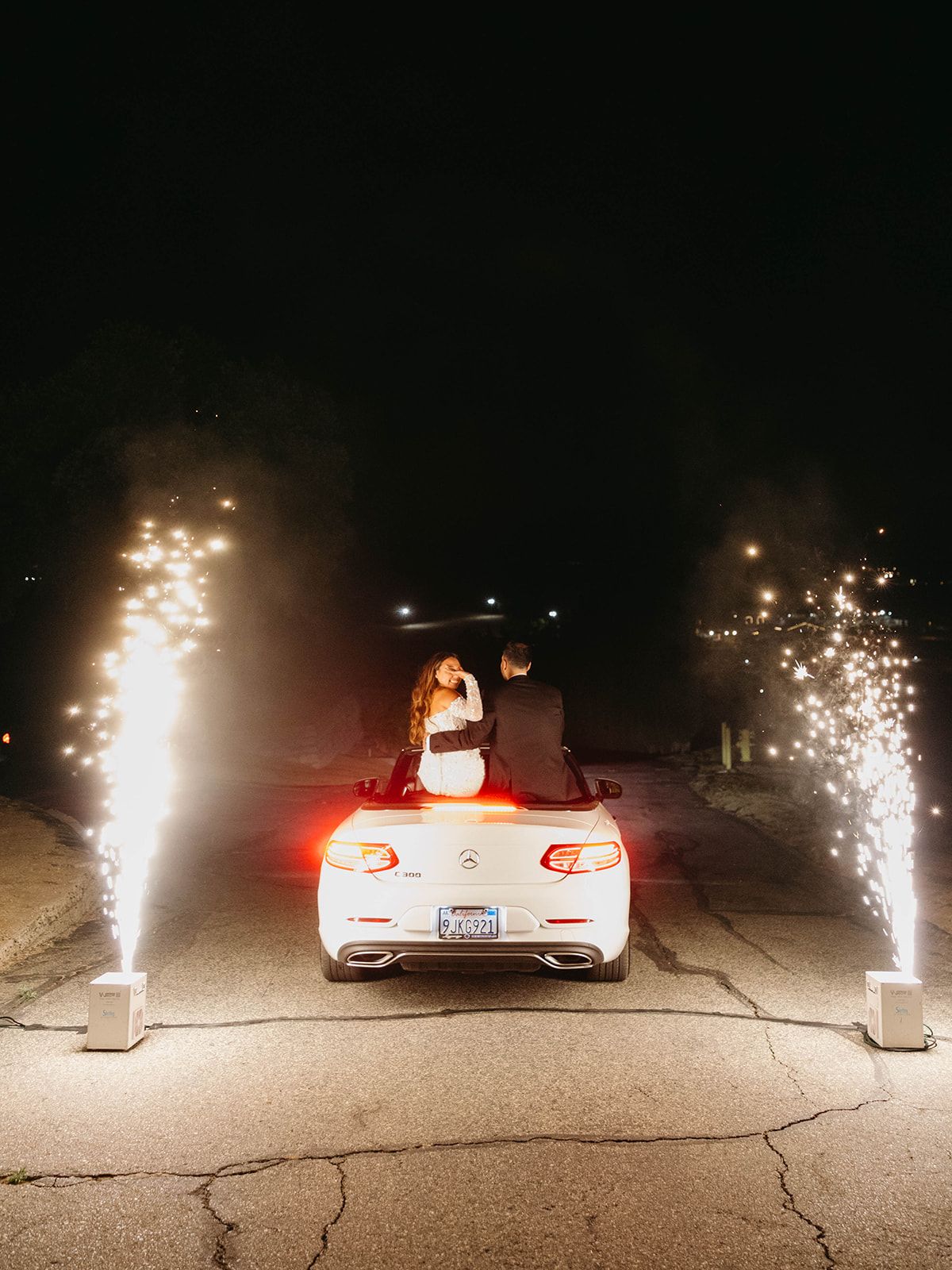 Wedding couple standing through car sunroof with sparklers creating a magical nighttime exit scene.