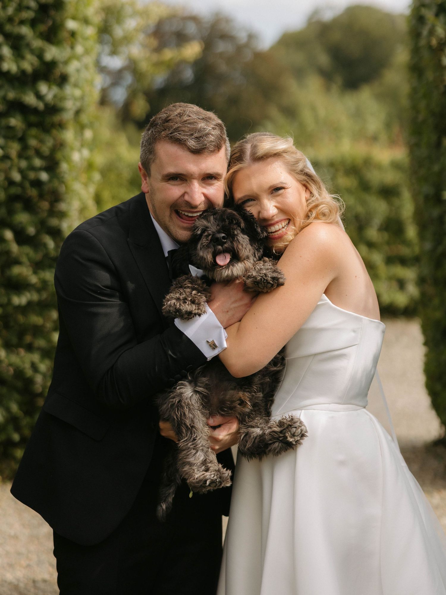 Wedding couple shares joyful moment with their fluffy brown dog during outdoor portraits.