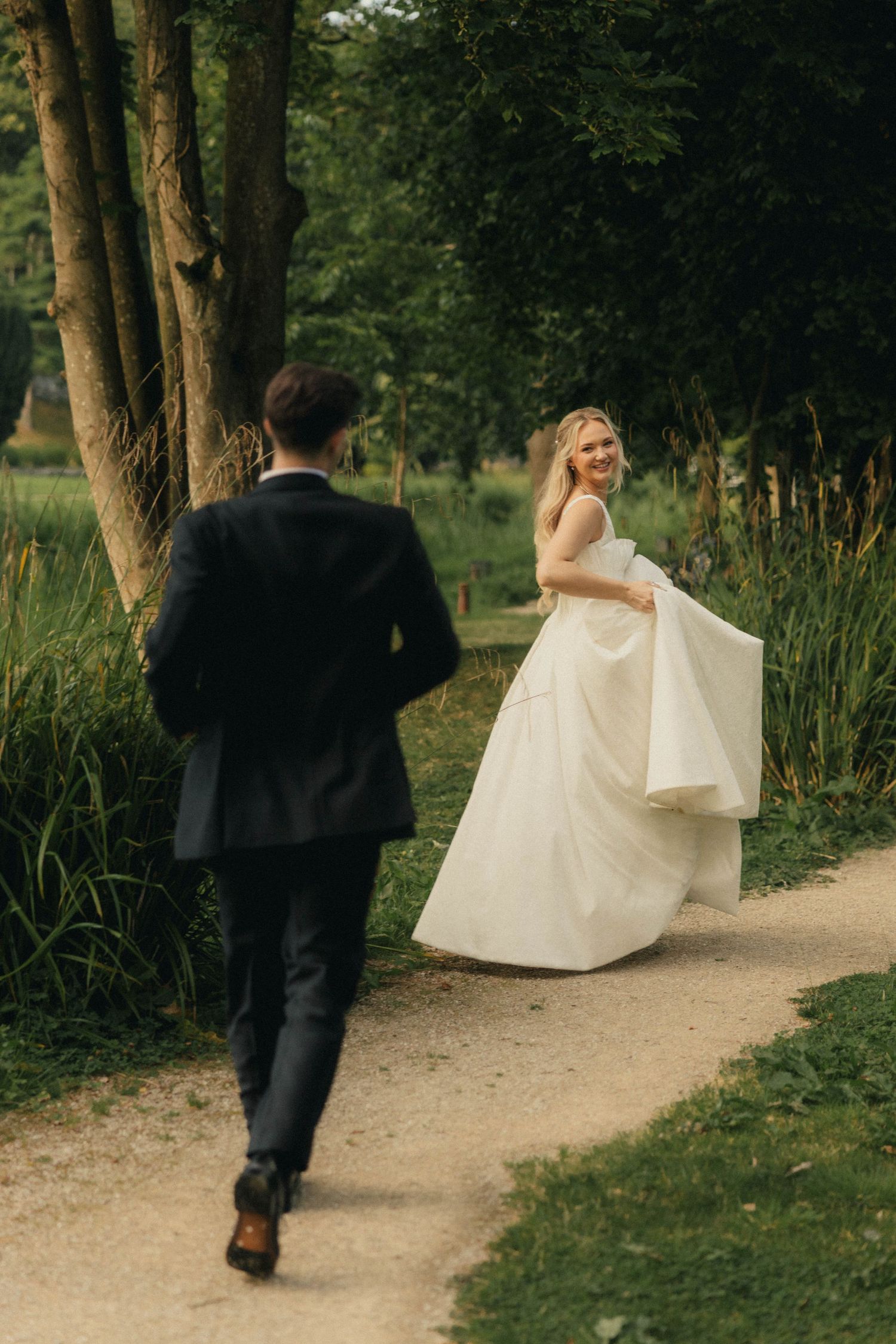 Bride in white gown smiles back at groom in black suit walking along a tree-lined garden path.