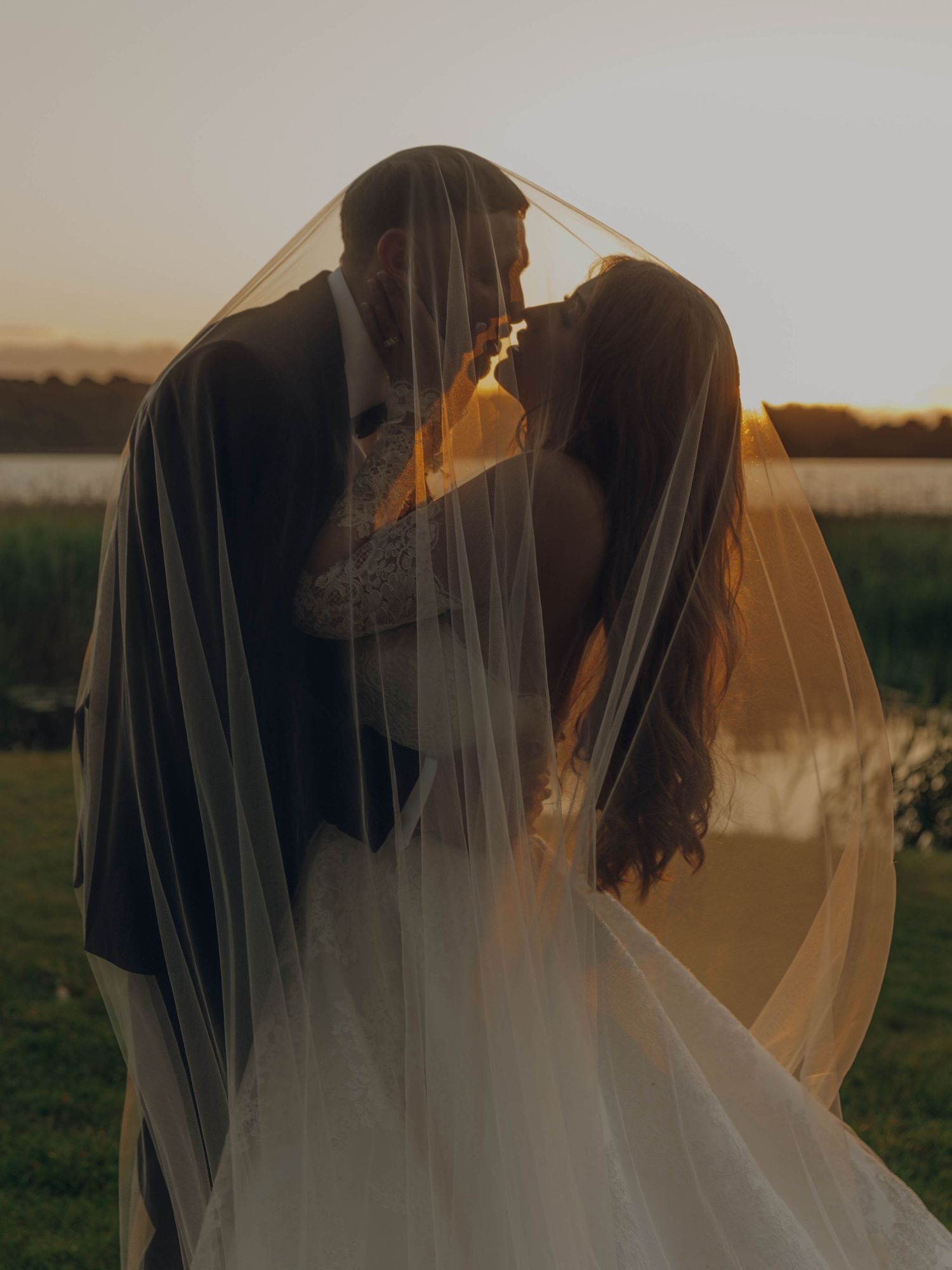 A romantic silhouette of a couple sharing an intimate moment under a flowing veil at sunset by a lake.