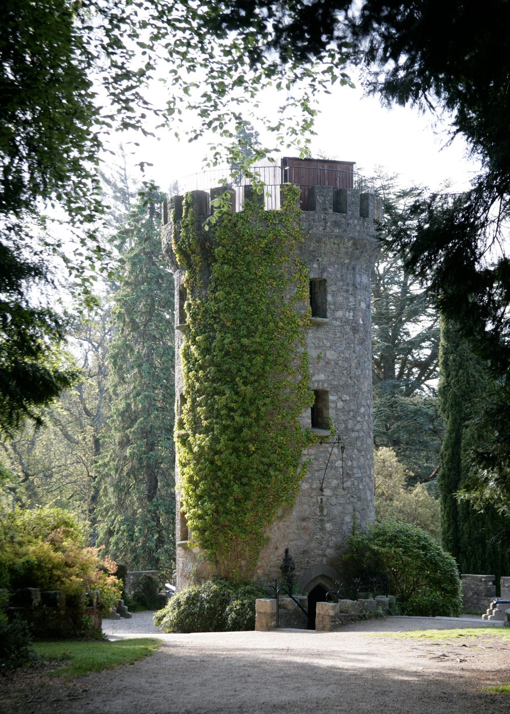 A medieval stone tower with windows stands among trees and greenery in a scenic outdoor setting.
