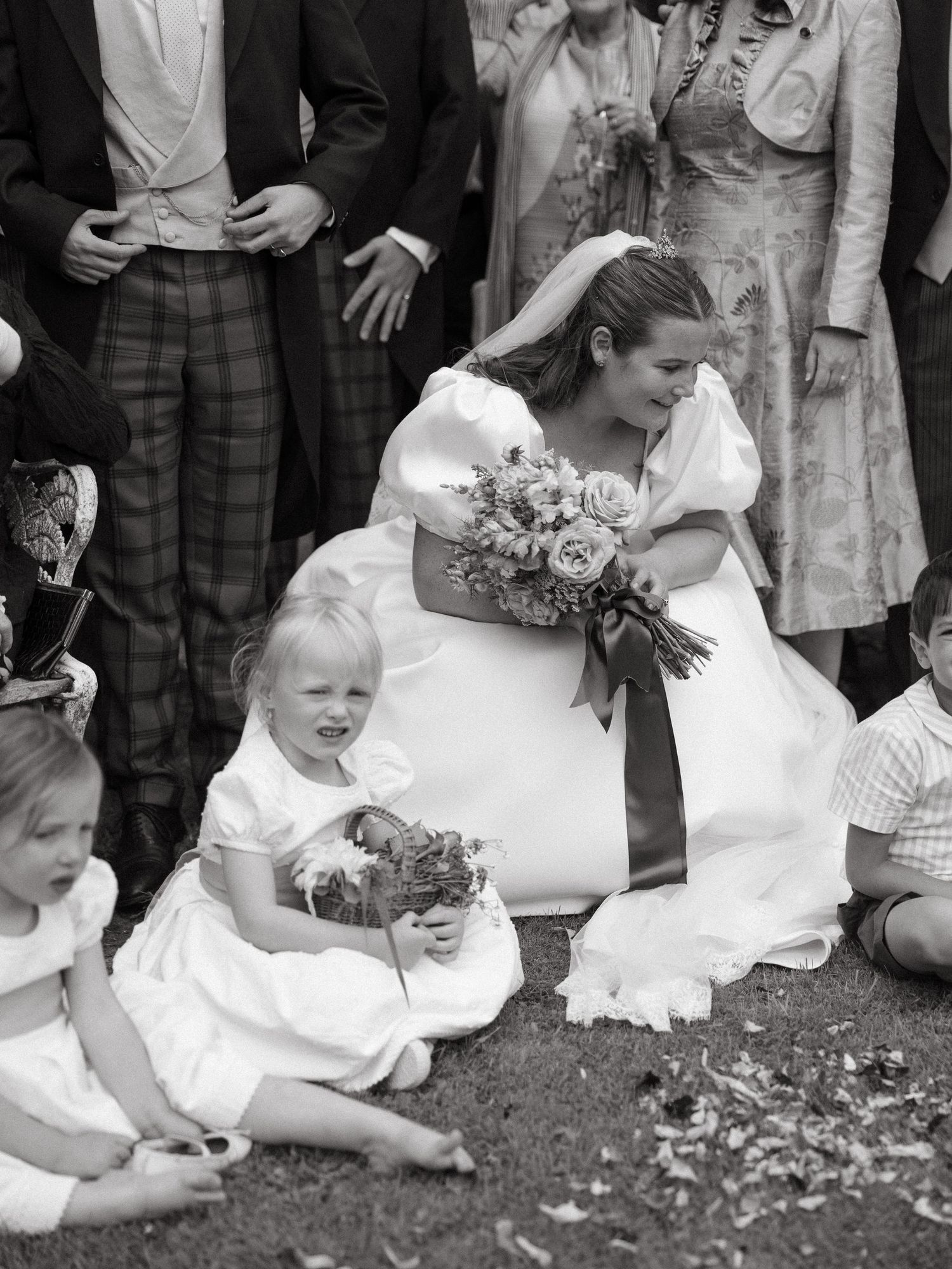 A bride in a white wedding dress crouches down and laughs while playing with a small child on the grass.