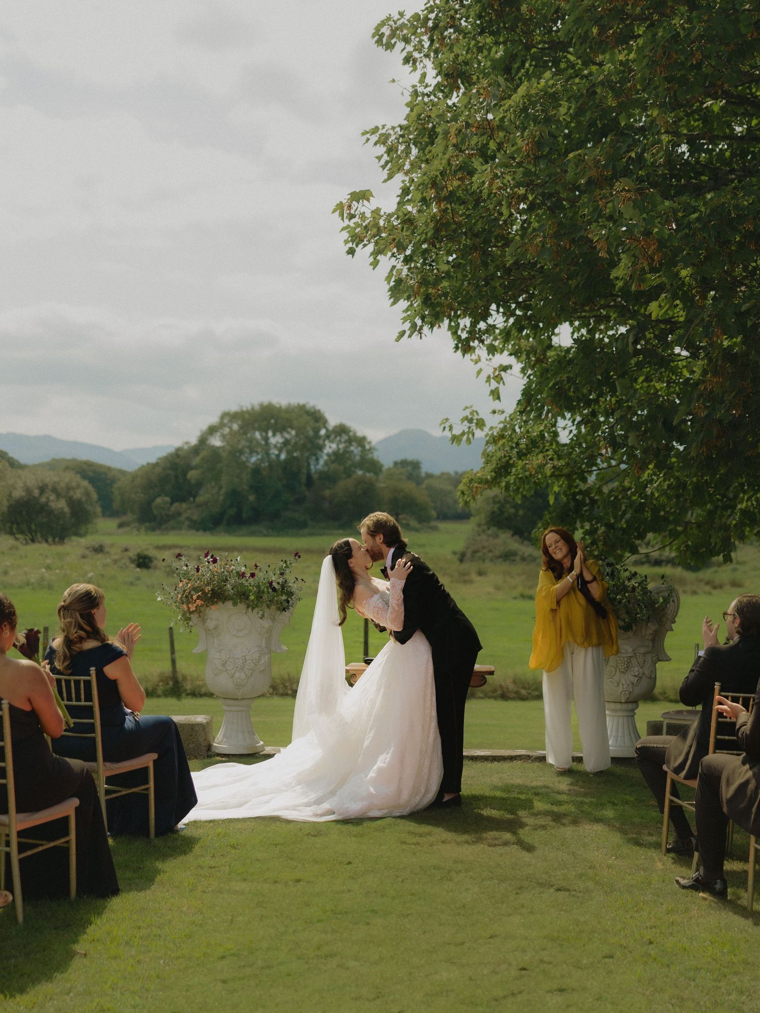 A wedding ceremony takes place outdoors on a grassy hillside with mountains in the background and guests seated under trees.