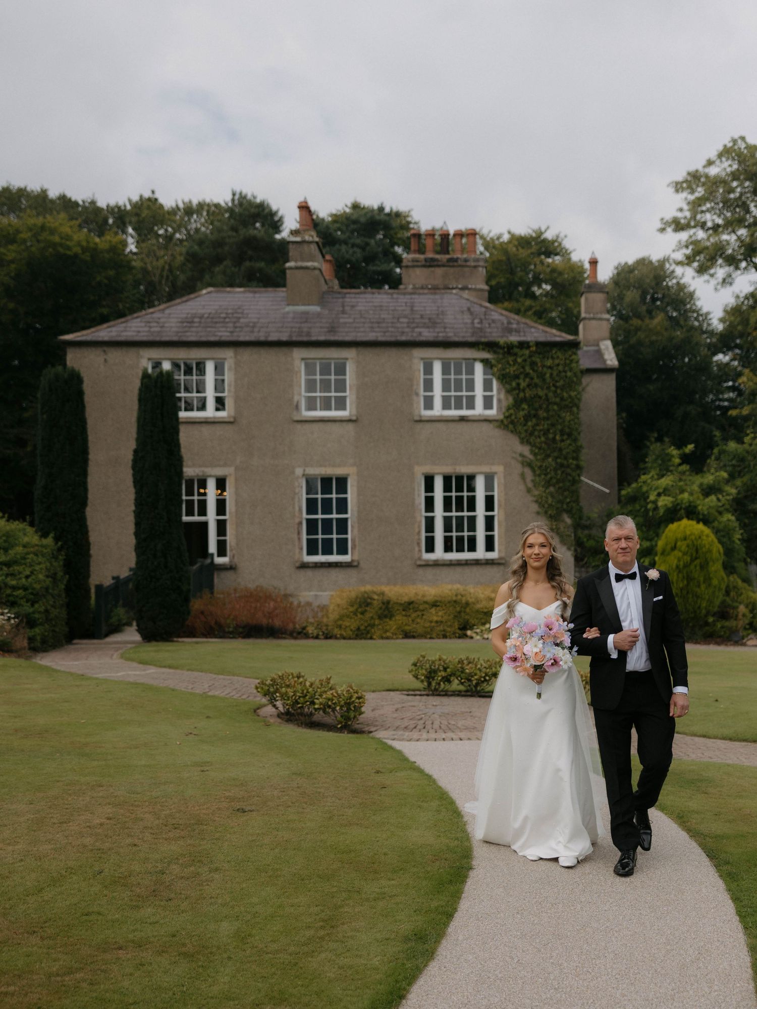 A couple walks along a curved driveway leading to an elegant stone country house on a landscaped estate.