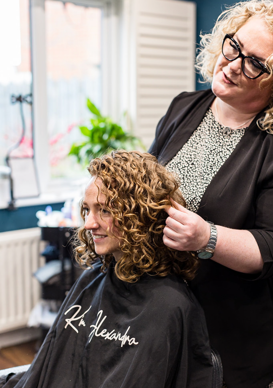 A hairstylist works on styling curly hair in a modern salon with natural lighting and indoor plants.