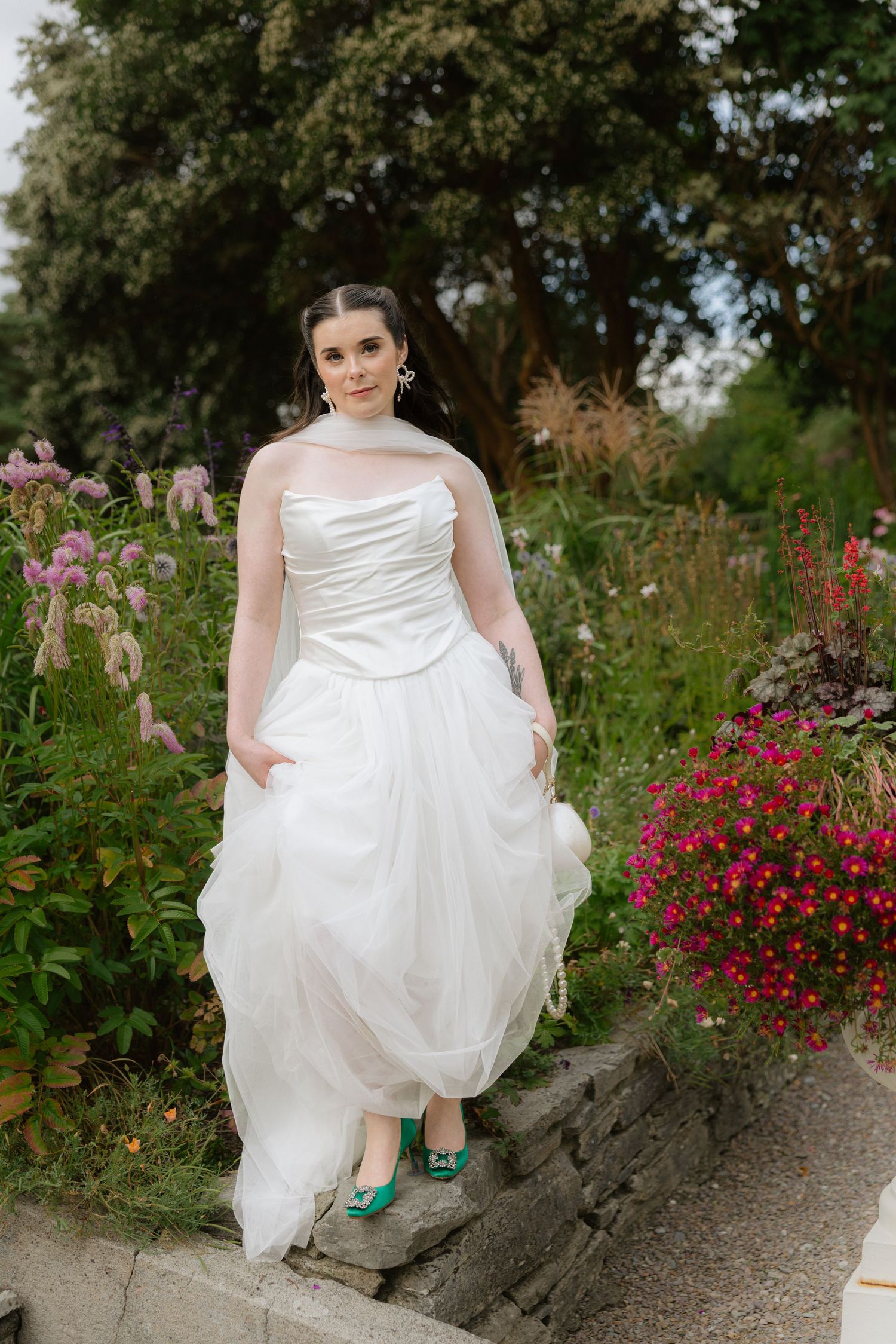 Bride in white strapless gown with tulle skirt and teal heels poses in lush garden with colorful flowers.