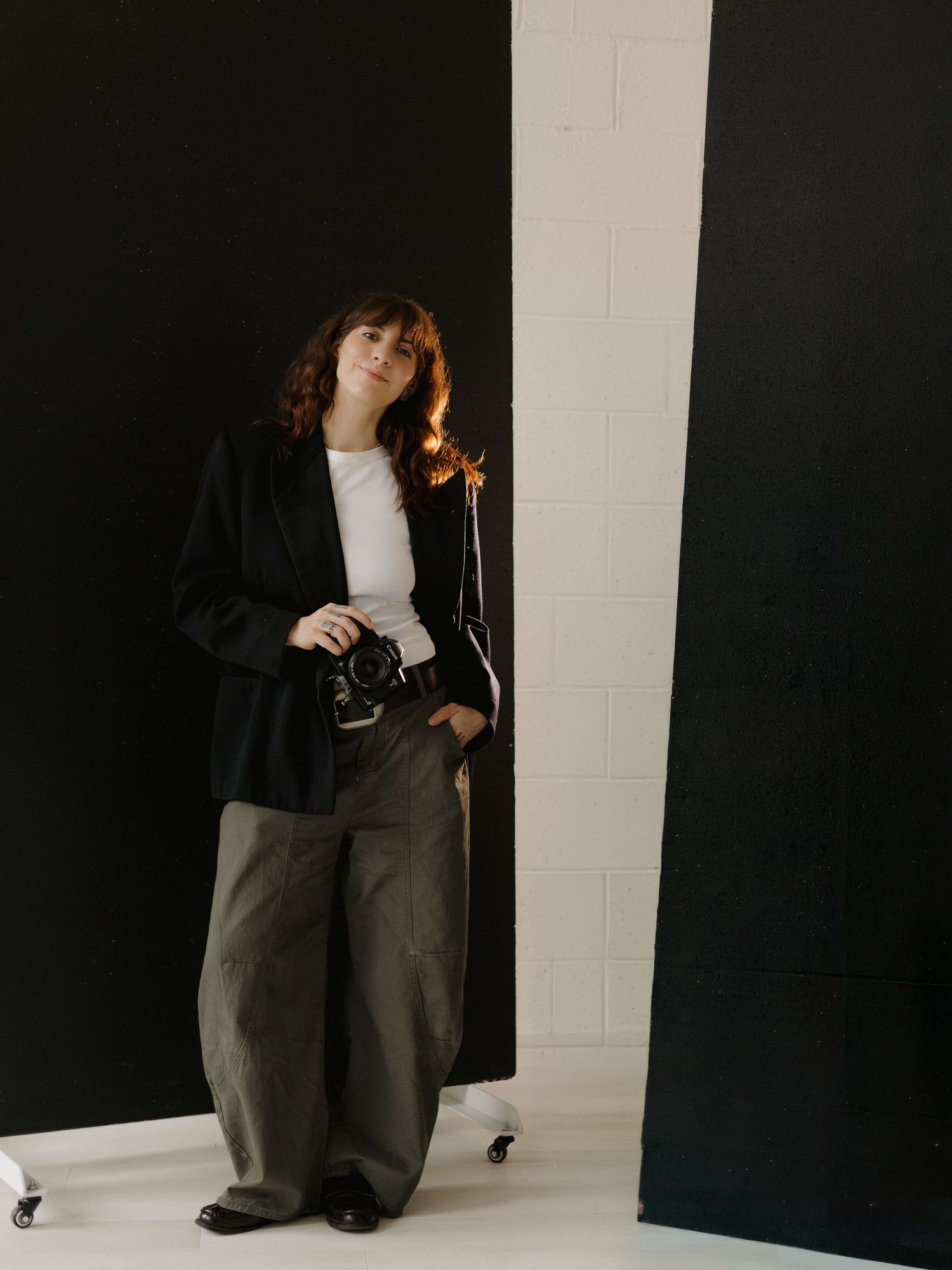 Woman in black blazer, white top, and olive wide-leg pants holds camera while posing against black and white backdrop.