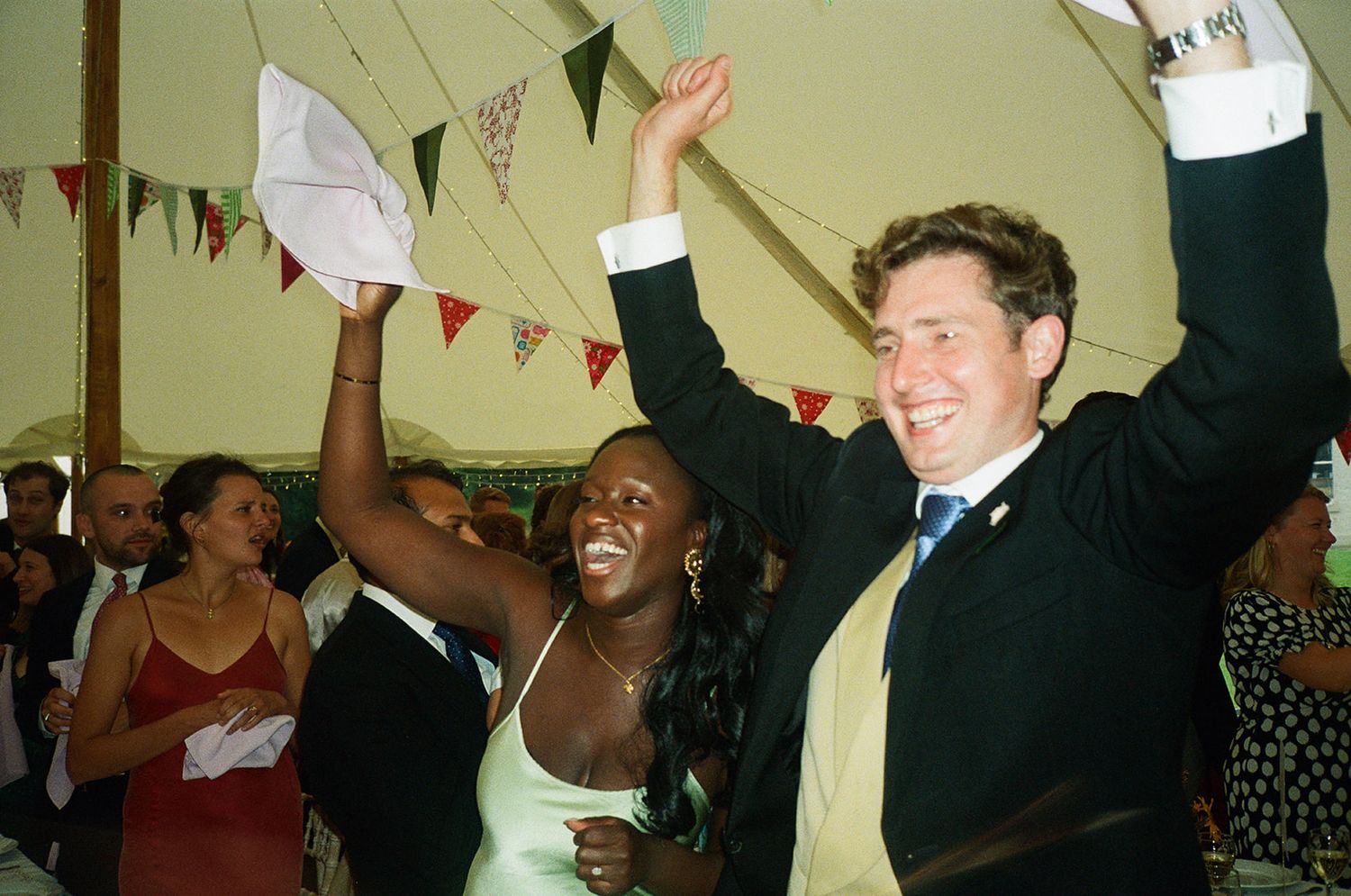 A group of people celebrating at a party with raised arms and joyful expressions under festive bunting decorations.
