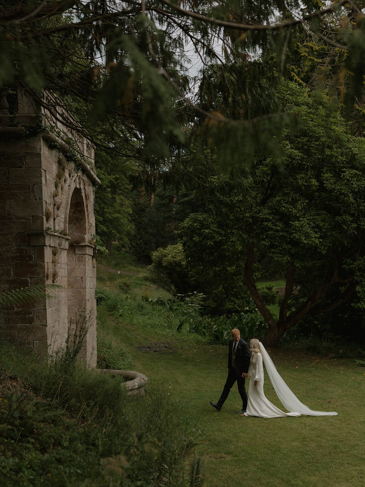A couple shares an intimate moment under a stone archway in a lush garden setting.
