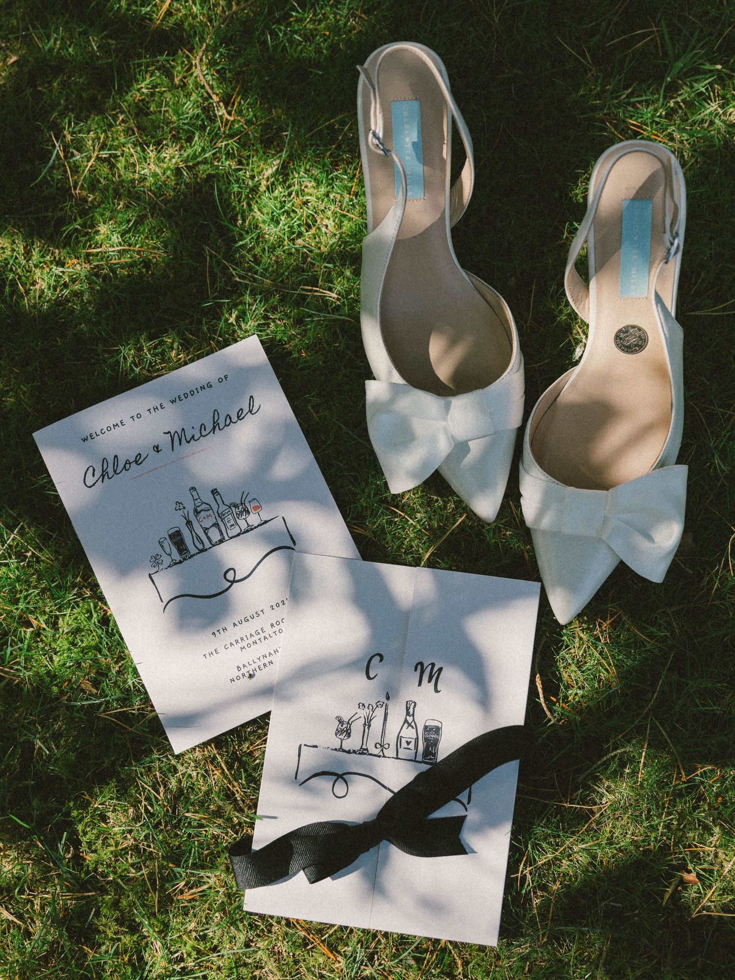 Wedding invitation cards and white bow heels arranged on grass in natural lighting.