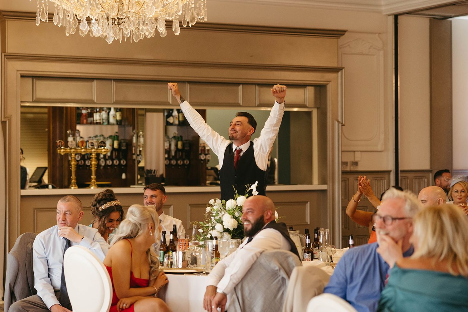 A wedding party celebrates at a reception with raised arms while a server stands victoriously on a chair in the background.
