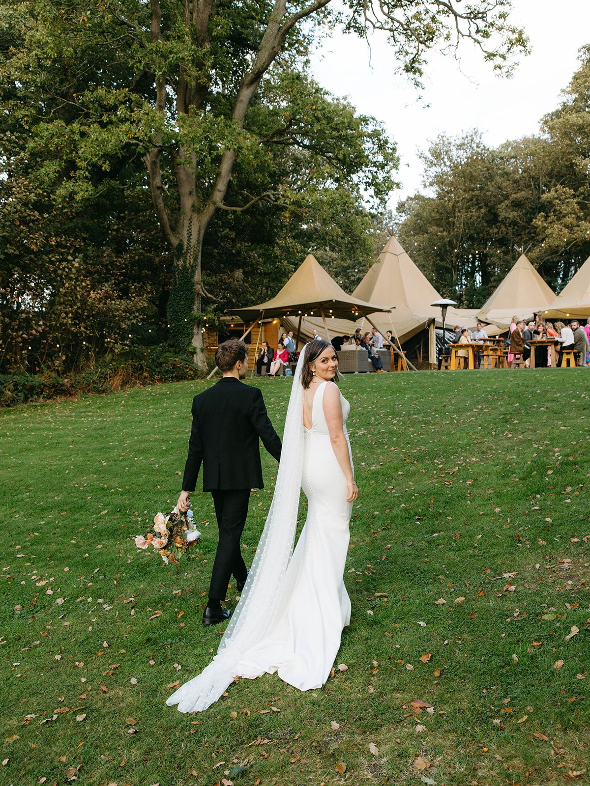 A couple walks across a green lawn towards tipi tents at an outdoor wedding reception.