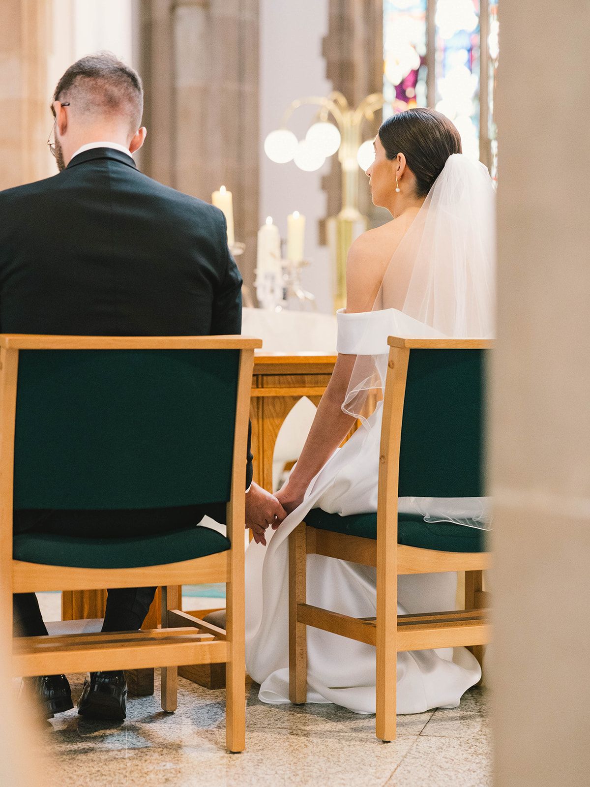 Bride and groom sit in green chairs during church wedding ceremony holding hands at the altar.