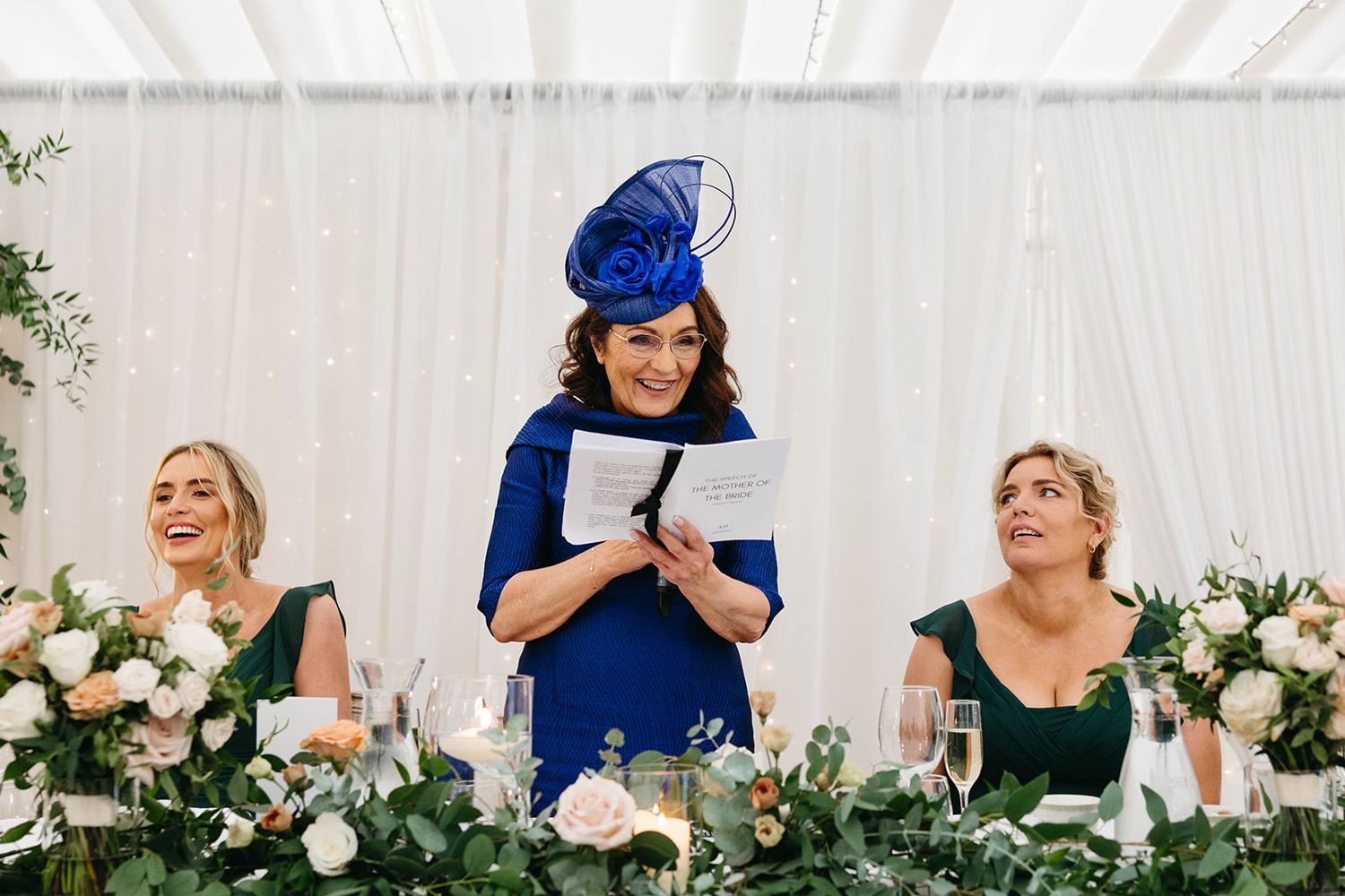 Wedding party members in elegant green and blue dresses smile while seated at floral-decorated reception table.