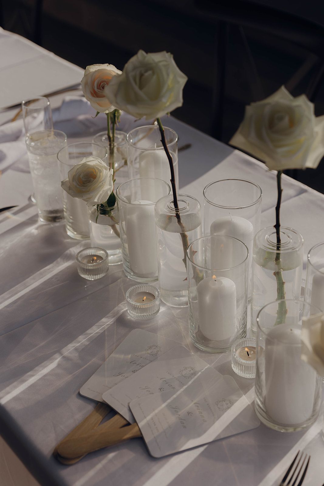 White roses in glass bud vases line a formal table setting with white candles and linens.