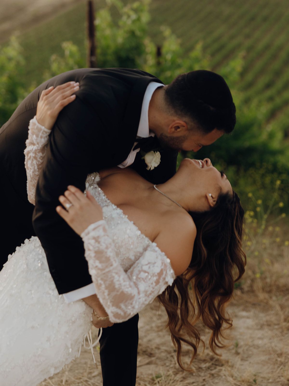 Romantic newlywed couple shares an intimate dip kiss amidst a lush green vineyard at sunset.