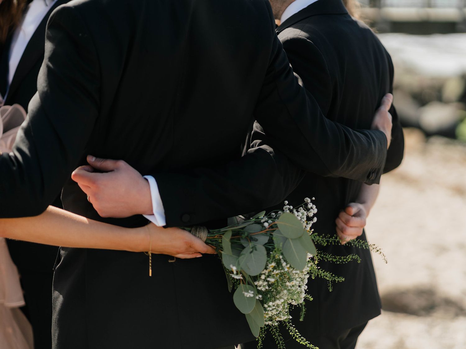 A close-up shot of two people embracing in black formal attire with a delicate bouquet of white flowers and eucalyptus.