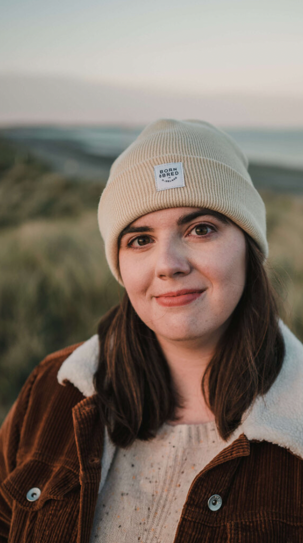 Young woman wearing a cream beanie hat and brown corduroy jacket smiling outdoors at golden hour.