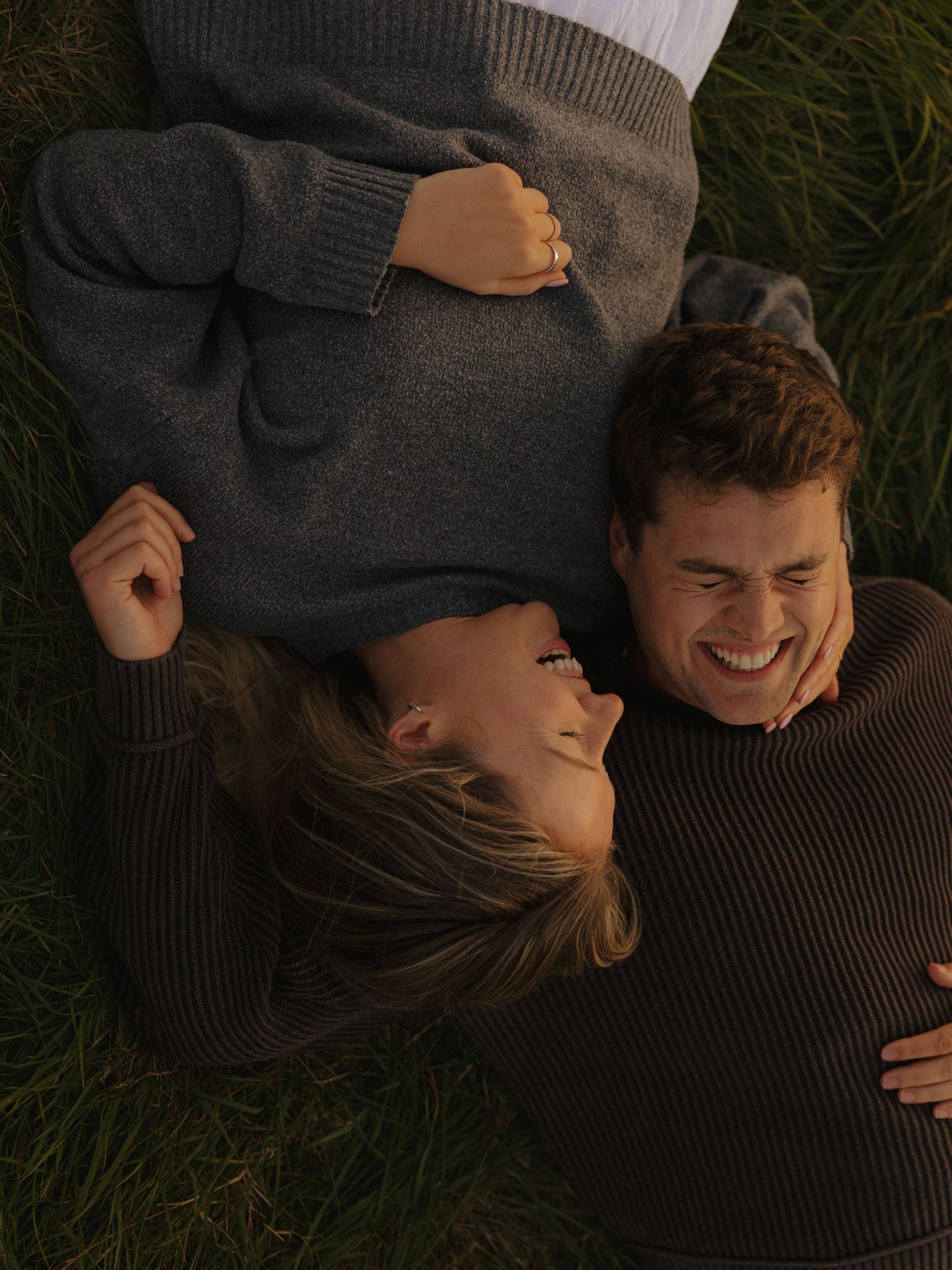 A couple lying on grass sharing laughter while wearing cozy sweaters.