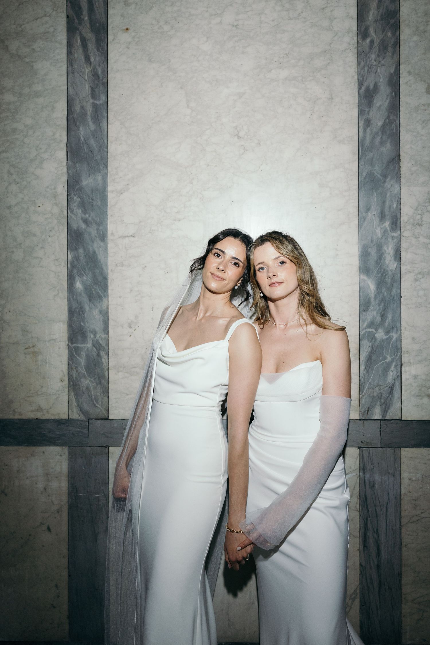 Two brides in elegant white gowns holding hands, one wearing a veil, posing together against a marble wall.