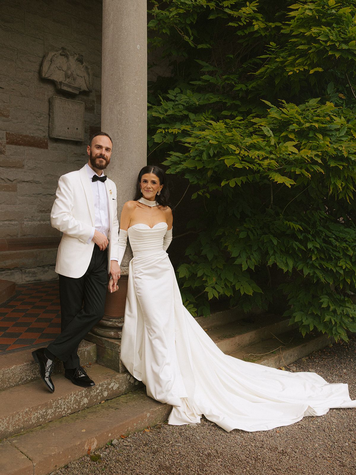 Elegant wedding couple poses on stone steps surrounded by lush greenery.