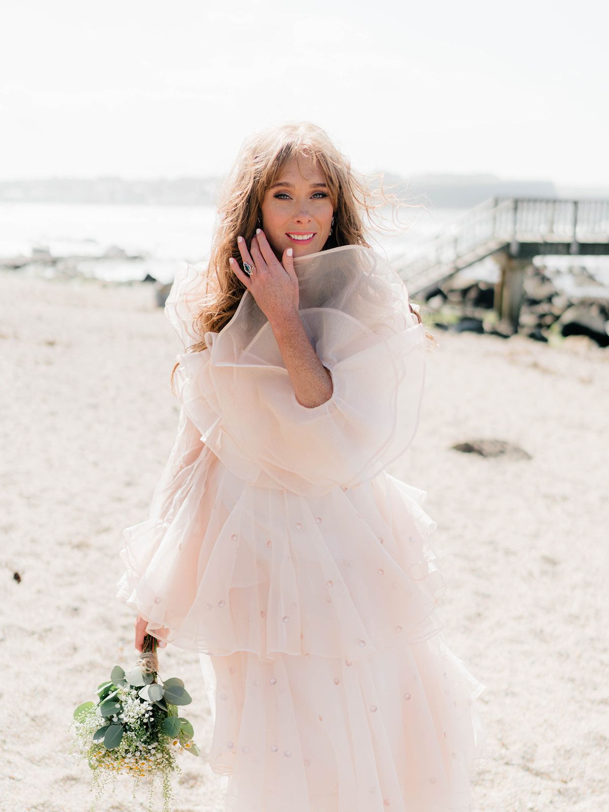 Joyful bridal portrait on a beach with soft pink ruffled dress and small bouquet.