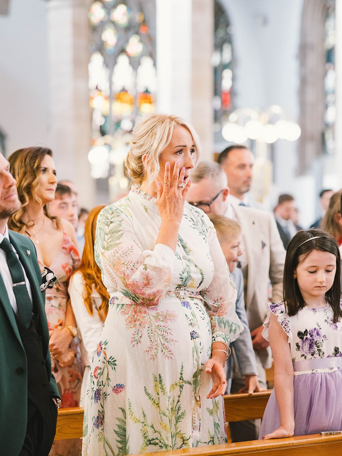 Wedding guests look on with emotion during a church ceremony beneath ornate stained glass windows and gothic architecture.