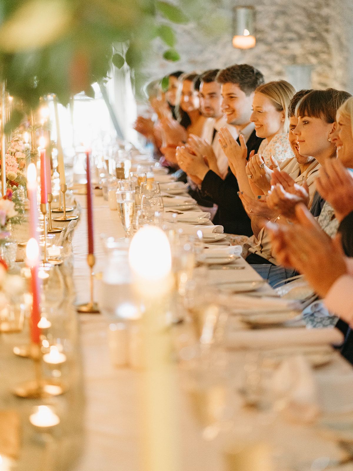 Wedding guests seated at an elegant candlelit dinner table clap and celebrate during an evening reception celebration.