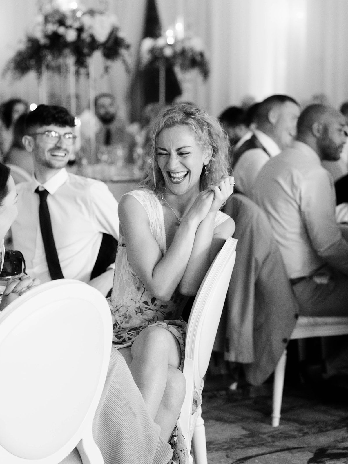 A candid black and white moment at a wedding reception shows guests laughing and enjoying themselves at decorated tables.