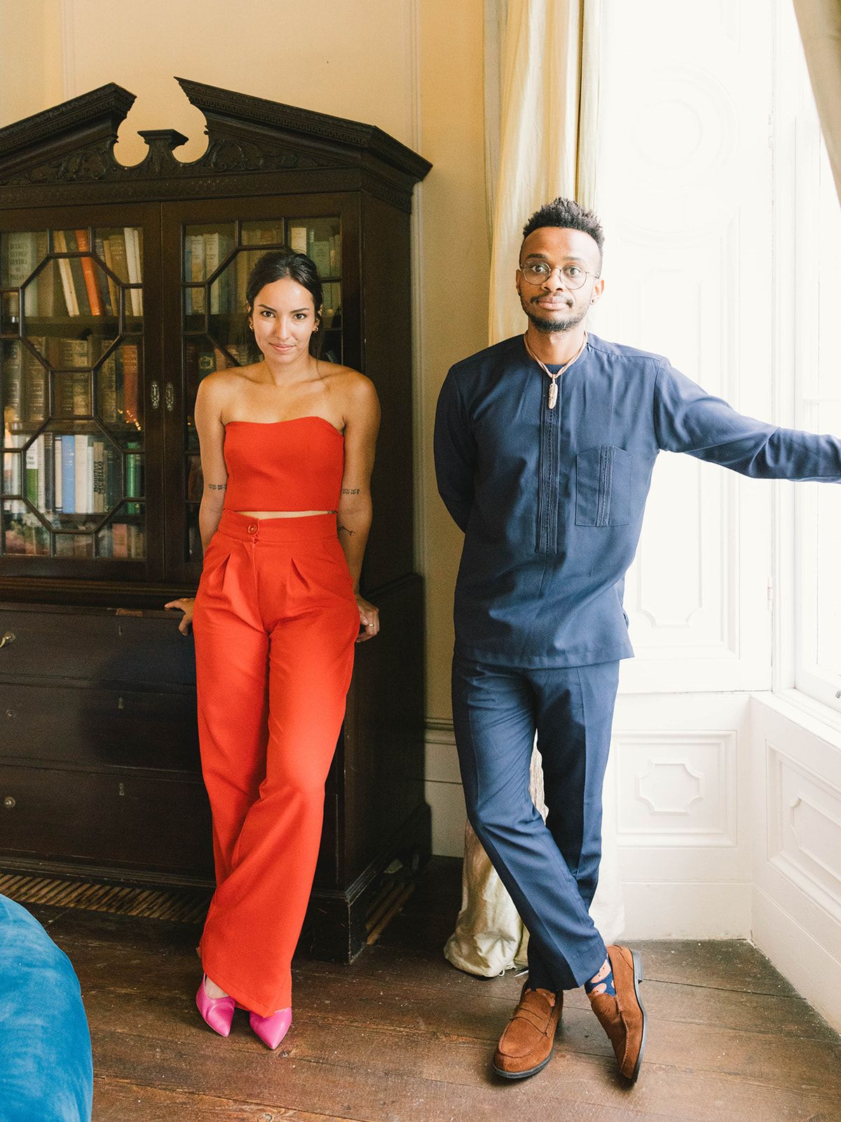 A couple poses in front of an antique display cabinet, with one person wearing a red jumpsuit and the other in blue denim.