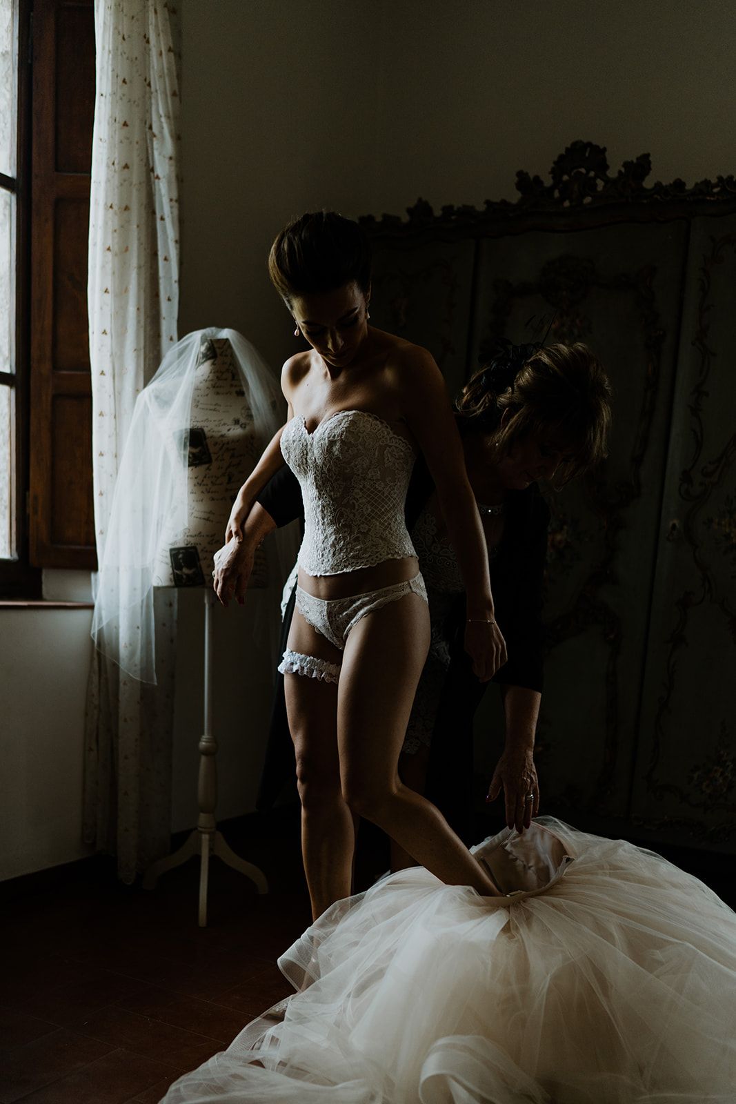 A moody boudoir photo shows someone getting dressed in white lingerie and wedding attire near a window.