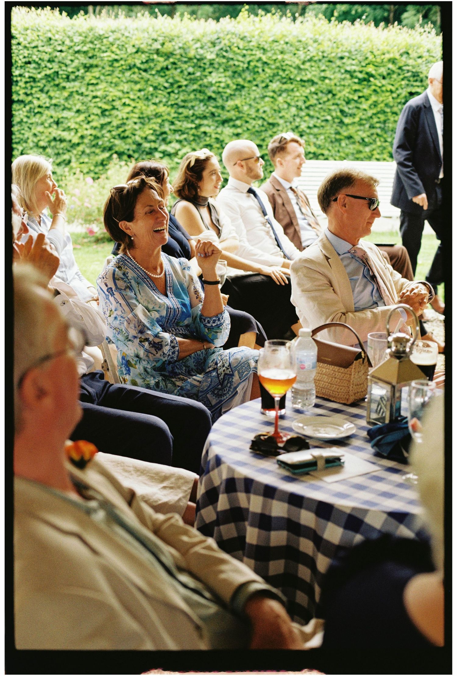 Wedding guests laughing and enjoying drinks at outdoor garden reception.
