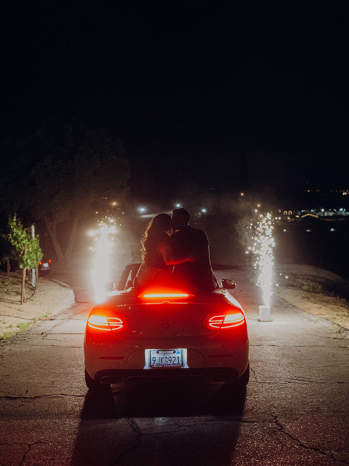Silhouettes sit on trunk of red Porsche car with illuminated taillights at night overlooking city lights in distance.