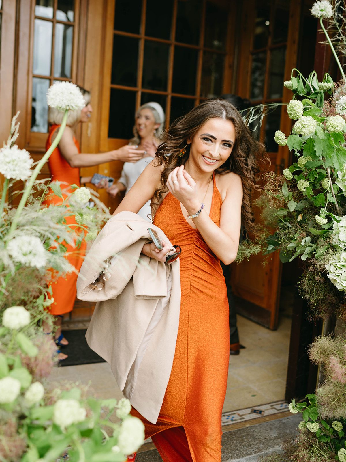 Bridesmaid in vibrant orange dress poses by floral-adorned doorway while holding a beige coat at outdoor event.