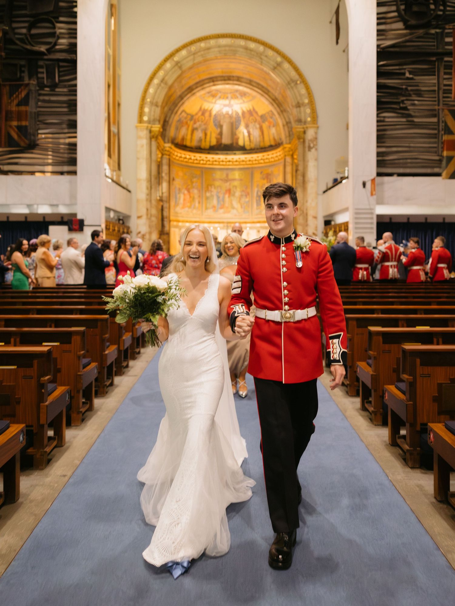Bride in white dress and British Royal Guard walking down blue-carpeted church aisle with ornate golden altar behind them.
