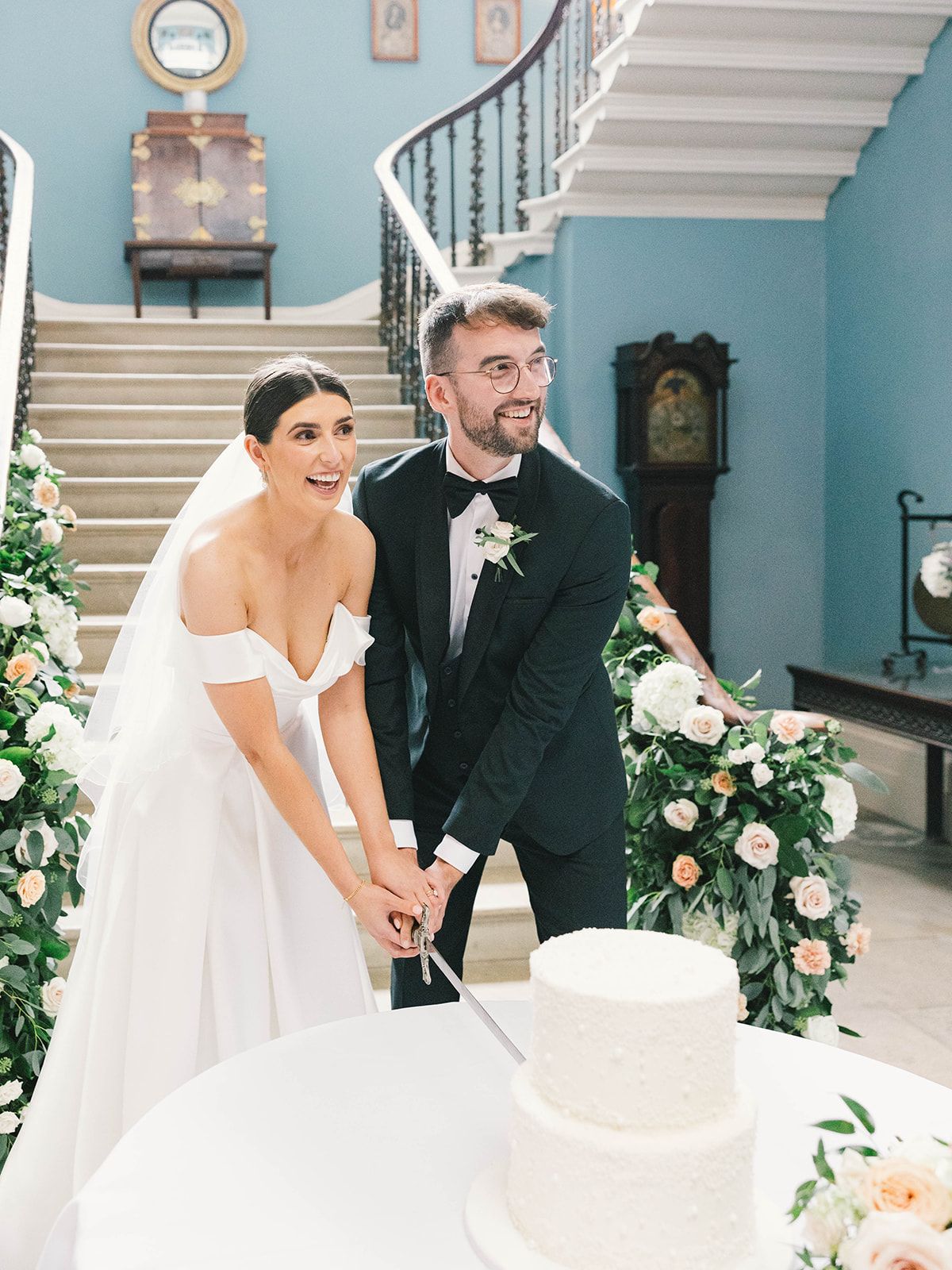 A wedding couple laughing together while cutting their white wedding cake in an elegant blue-walled staircase setting.