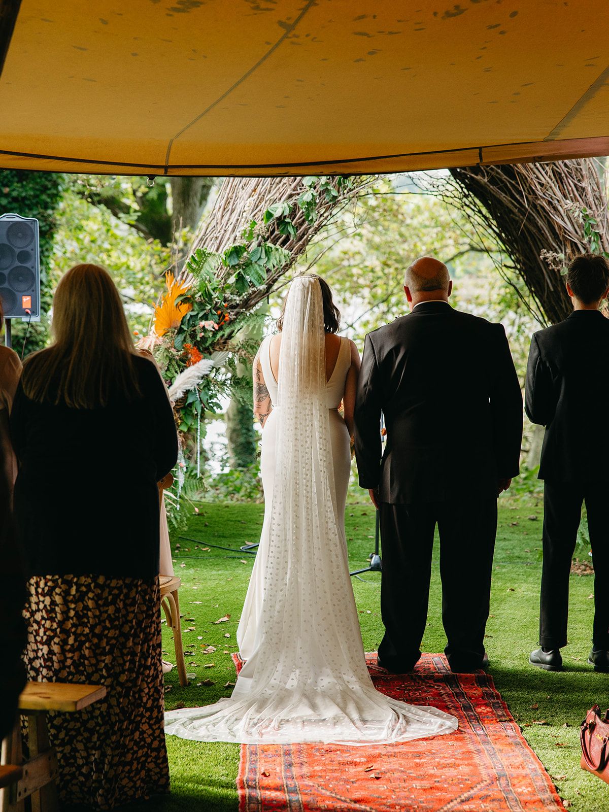 Wedding ceremony taking place under a rustic outdoor canopy with a long white wedding dress train on a red carpet.