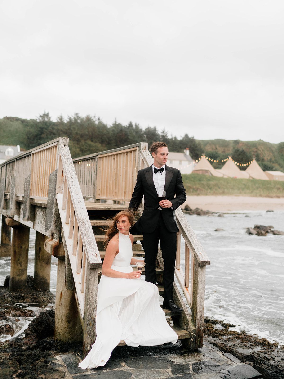 A couple poses on a wooden boardwalk by the ocean with teepee tents visible in the background.