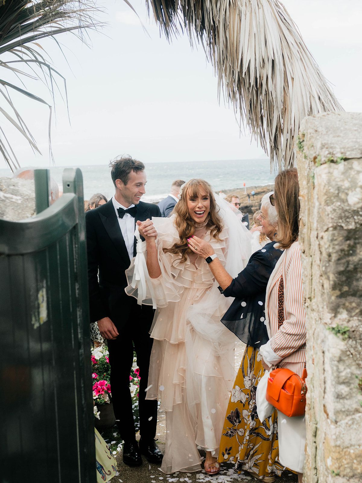 Wedding couple sharing joyful moment under palm fronds by seaside gate.