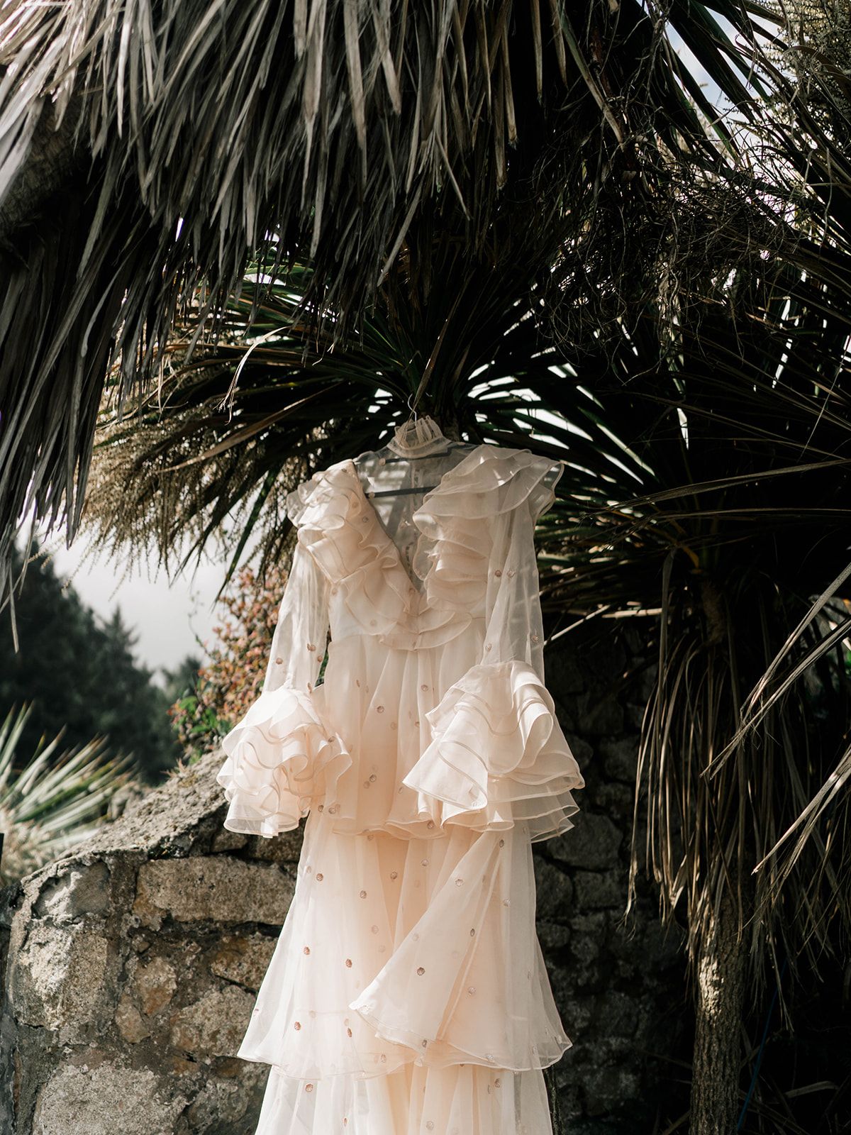 Ethereal peach-colored wedding dress with ruffled details hangs against tropical palm fronds outdoors.