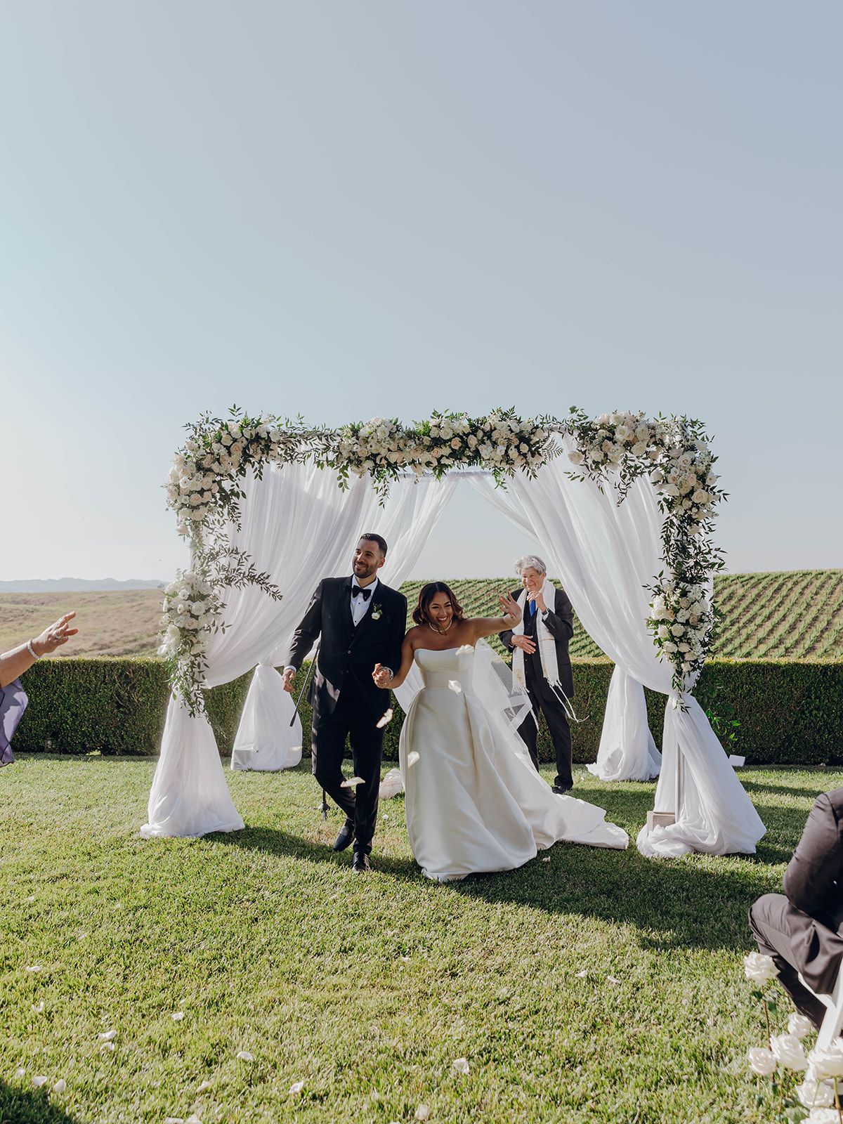A joyous wedding celebration beneath an elegant floral arch with white curtains against rolling vineyard hills.