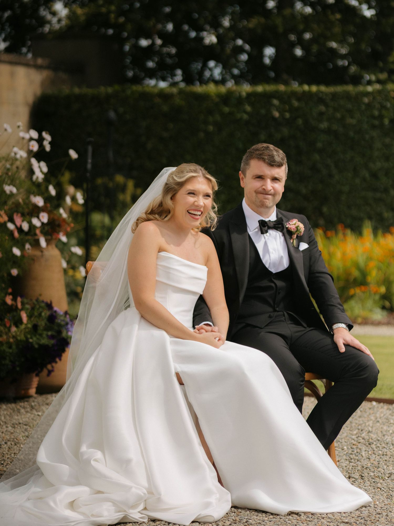 Couple in wedding attire sitting together outdoors with flower petals falling around them.