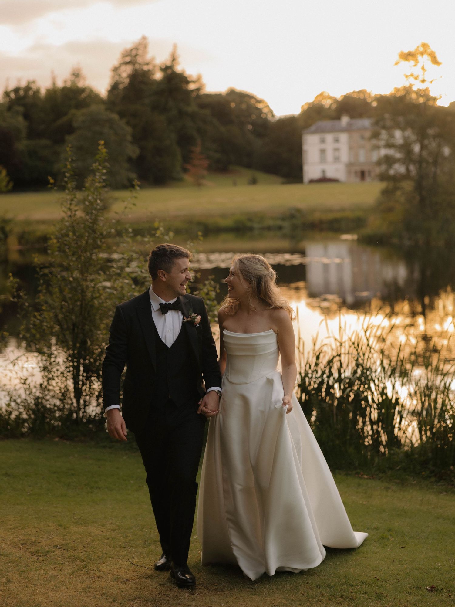 Golden hour wedding portraits by a lake with couple walking through lush grounds.