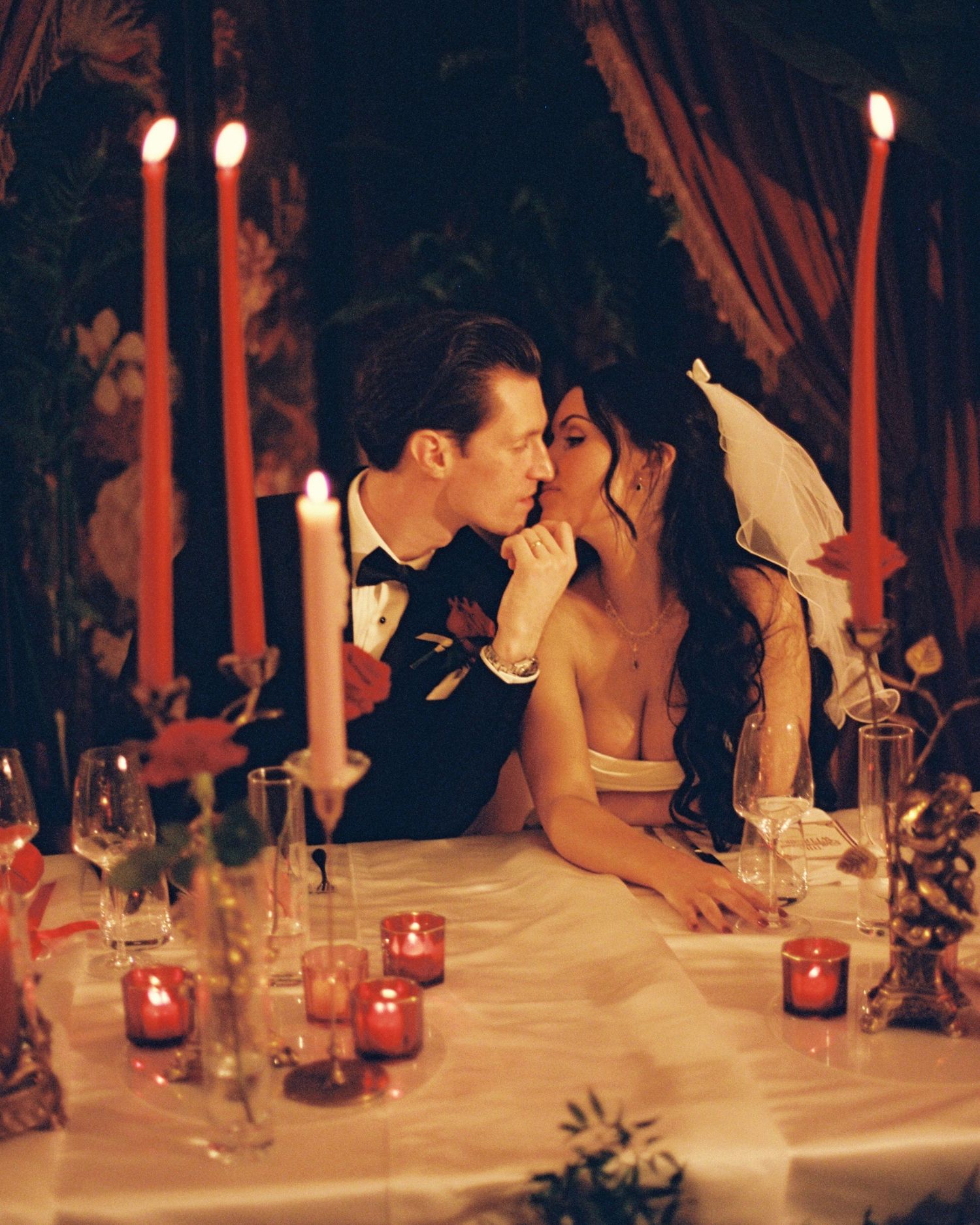 Bride and groom share a romantic candlelit moment at their wedding reception table adorned with red candles.