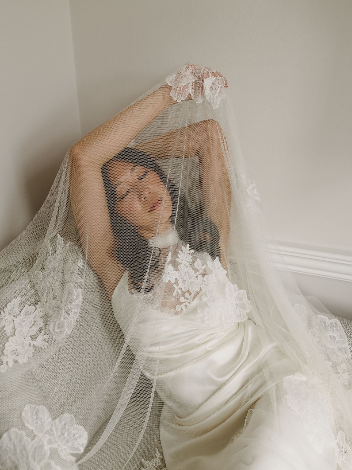 Bride in white satin gown poses gracefully under floral lace veil with eyes closed in soft natural light.