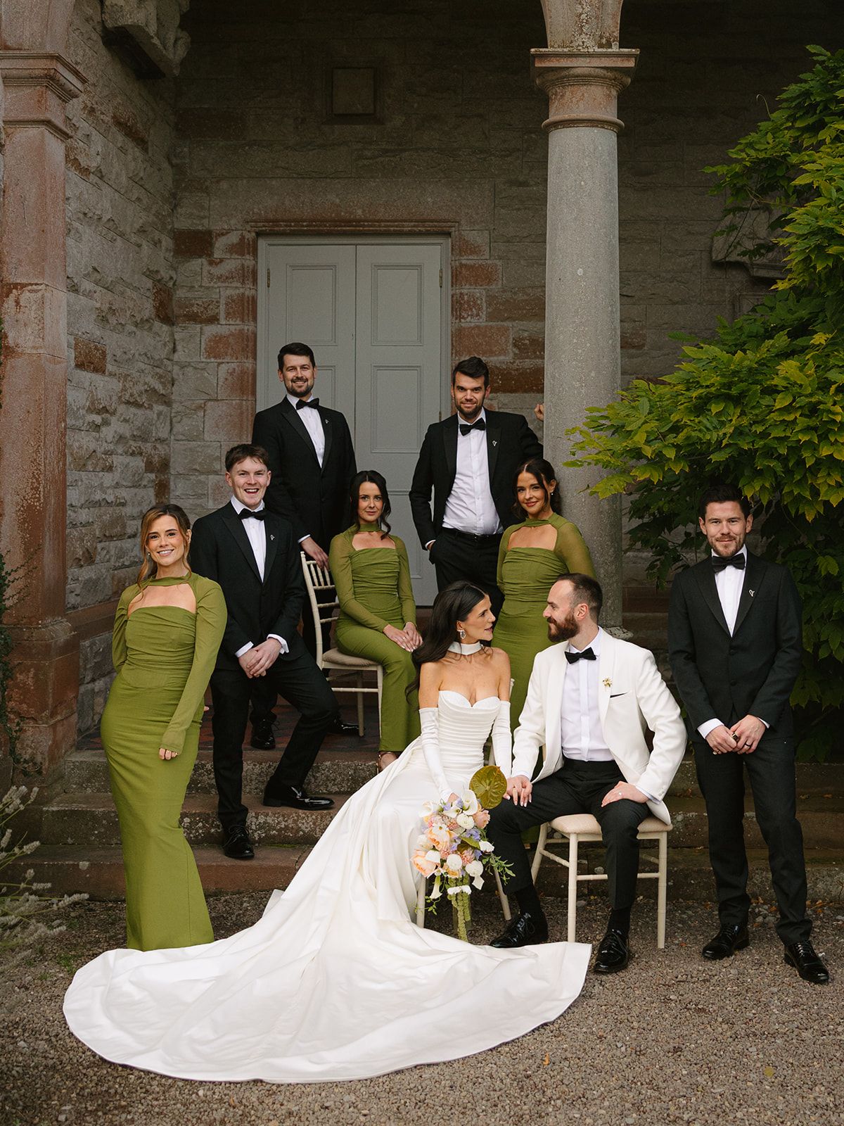 Elegant wedding party poses on stone steps with bridesmaids in olive green dresses and groomsmen in black tuxedos.