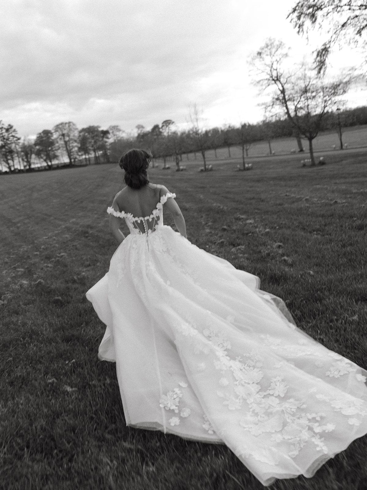 A flowing white wedding dress train billows dramatically across a grassy field in black and white photography.